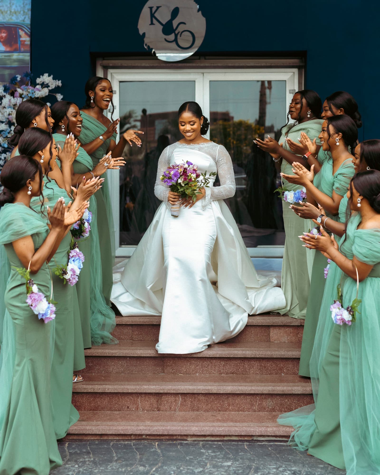 Kechi descends the steps in her white gown as her bridesmaids applaud.